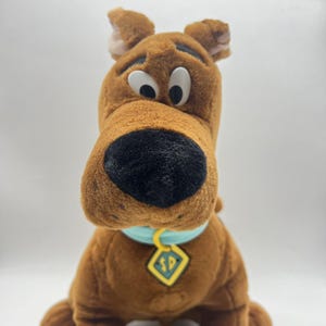 May include: A plush toy of Scooby-Doo, a brown dog with a large black nose, white eyes, and a blue collar with a yellow tag. The toy is made of soft, fuzzy material and has a friendly expression.