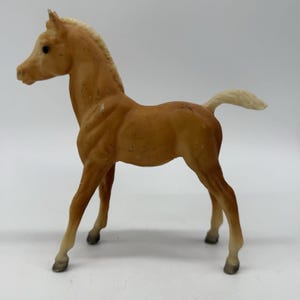 Op de afbeelding: Een bruin speelgoed paardenfiguur met een lichtgekleurde manen en staart. Het paard staat met gespreide benen en zijn hoofd is iets naar links gedraaid. Het speelgoed is gemaakt van een stevig materiaal en heeft een glad oppervlak.