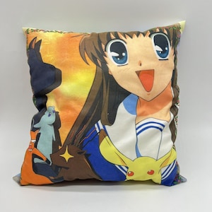 May include: A colorful pillow featuring an anime character with long brown hair, wearing a blue and white sailor uniform. The character is smiling with her mouth open, and her eyes are wide open. The pillow also features a small yellow and white cartoon character, a blue and white cartoon character, and a red and white cartoon character.