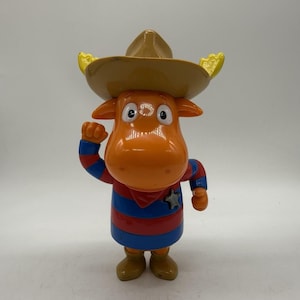 Puede incluir: Figura de juguete de plástico de un alce de dibujos animados con un sombrero de vaquero marrón con detalles amarillos. El alce es naranja con una camisa a rayas azules y rojas y botas marrones. Está saludando con la mano derecha.