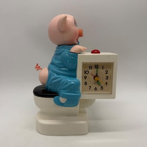 May include: A novelty analog clock featuring a pink pig figure wearing blue pajamas, sitting on a white toilet with a black seat. The clock face is square, white with black numerals, and has green hour and minute hands with a yellow second hand.