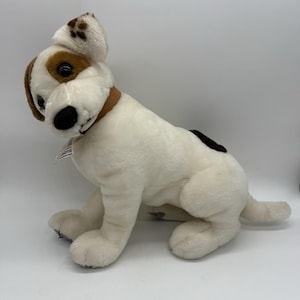 Puede incluir: Un peluche de perro Jack Russell Terrier. El peluche es blanco con marcas marrones en las orejas y alrededor de los ojos. Tiene un collar marrón y una mancha negra en la espalda. El perro está sentado.