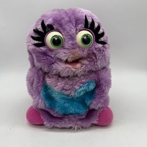 May include: A plush toy featuring purple shaggy fur, large green eyes with prominent black eyelashes, and a blue patch on the chest. The creature has a small open mouth showing a pink interior and two small pink feet at the base of the body.