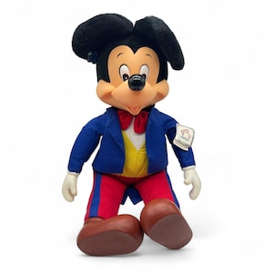 Peut inclure: Une peluche de Mickey Mouse portant un costume bleu avec un gilet jaune et un pantalon rouge. La peluche a un n&oelig;ud papillon rouge et une &eacute;tiquette blanche sur le bras qui dit "Disneyland".