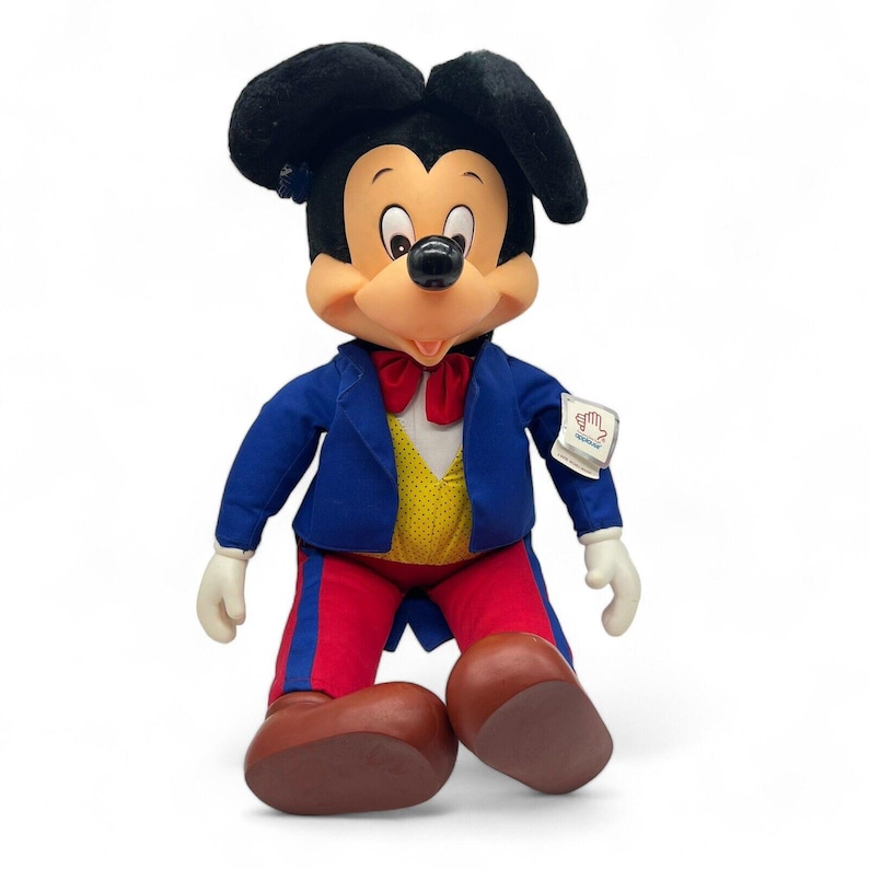 Peut inclure: Une peluche de Mickey Mouse portant un costume bleu avec un gilet jaune et un pantalon rouge. La peluche a un n&oelig;ud papillon rouge et une &eacute;tiquette blanche sur le bras qui dit "Disneyland".