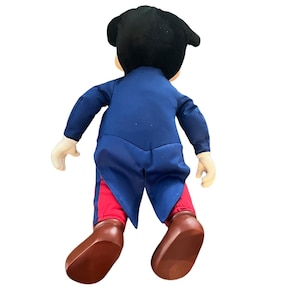 Peut inclure: Un jouet en peluche repr&eacute;sentant un personnage de dessin anim&eacute; portant un costume bleu avec des accents rouges et des chaussures noires. Le jouet est debout sur un fond blanc.