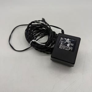 May include: A black Atari power adapter with a long, coiled black cord. The rectangular unit features a label with the Atari logo, part number C010472, and technical specifications including input 120V AC 60Hz 12W and output 9V DC 500mA, made in Taiwan.