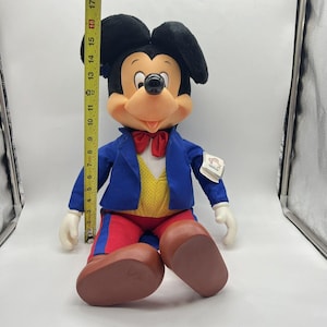 Peut inclure: Une peluche de Mickey Mouse portant un costume bleu avec un gilet jaune et un pantalon rouge. La peluche est assise les jambes crois&eacute;es et porte un n&oelig;ud papillon rouge. La peluche mesure environ 43 cm de haut.