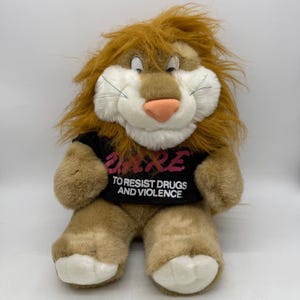 May include: A plush lion toy with tan fur, a shaggy orange mane, and white accents on the face and paws. The lion wears a black t-shirt featuring the pink text D.A.R.E. and the white text TO RESIST DRUGS AND VIOLENCE printed on the front.