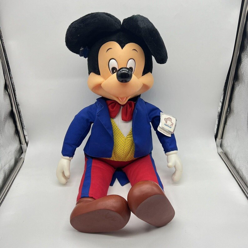 Peut inclure: Une peluche de Mickey Mouse portant un costume bleu avec un n&oelig;ud papillon rouge et un gilet jaune. La peluche est assise les jambes crois&eacute;es et porte une &eacute;tiquette sur le bras qui dit "Disney Store".