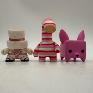 May include: Three colorful figurines. The first is a white figure with a hat and pink accents. The second is a striped red and pink figure. The third is a pink dog-like figure with large ears.