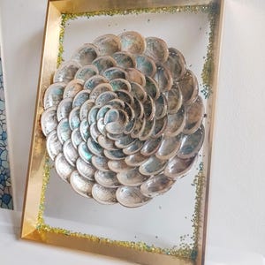 Op de afbeelding: Een ingelijst kunstwerk met een spiraalvormige opstelling van iriserende schelpen. De schelpen zijn geplaatst tegen een transparante achtergrond in een goudkleurige lijst, geaccentueerd met groene en gouden glitter.