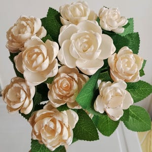 Peut inclure: Un bouquet de roses artificielles de couleur crème avec des feuilles vertes. Les roses ont plusieurs couches de pétales et les feuilles sont d'un vert profond. L'arrangement est étroitement regroupé, créant une apparence pleine et luxuriante.