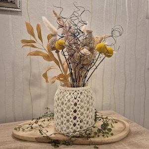 Könnte beinhalten: Eine dekorative Vase mit cremefarbenem Makramee-Überzug enthält ein Trockenblumenarrangement. Das Arrangement umfasst gelbe, braune und beige Elemente mit einigen schwarzen Drahtakzenten. Die Vase steht auf einem Holztablett mit Grün.