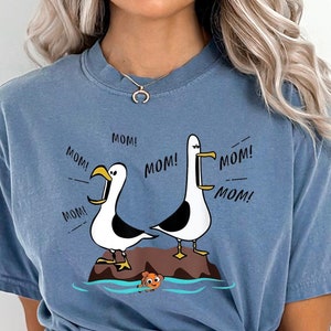 May include: A blue t-shirt with a graphic of two seagulls saying "Mom!" on a rock with a fish swimming in the water below. The text on the shirt is "Mom!" repeated multiple times.