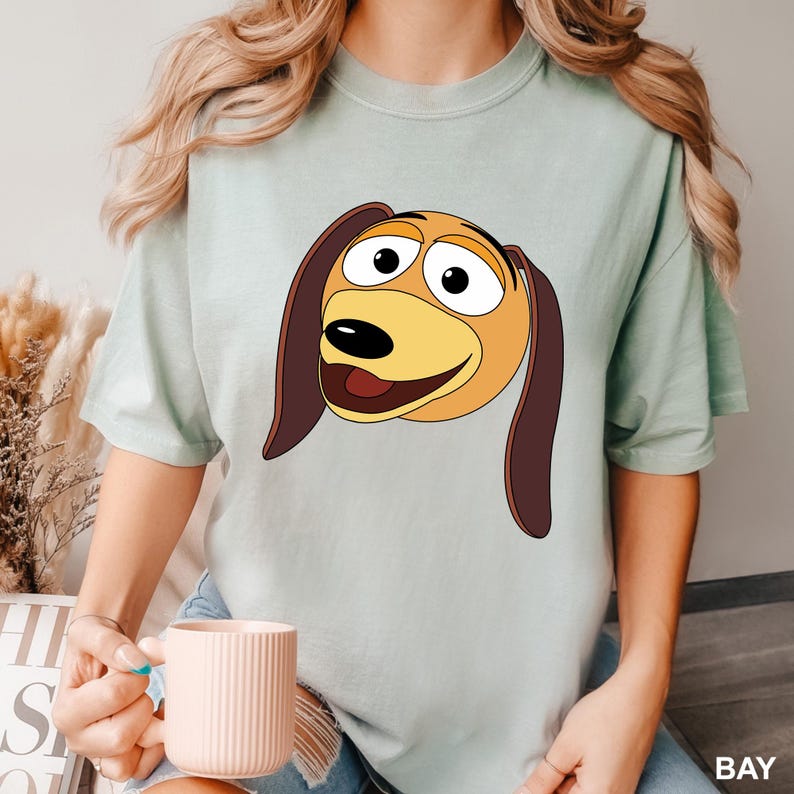 Puede incluir: Una camiseta azul claro con una imagen de dibujos animados de un perro marr&oacute;n y amarillo con ojos grandes y una amplia sonrisa. Las orejas del perro son ca&iacute;das y marrones.