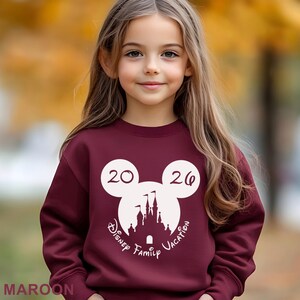A young girl with long blonde hair wearing a maroon sweater with a Disney-themed design and the text "Disney Family Vacation" printed on it.