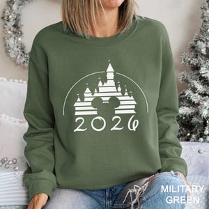 A woman wearing a green sweatshirt with a white graphic of a castle and the year "2026" printed on it.