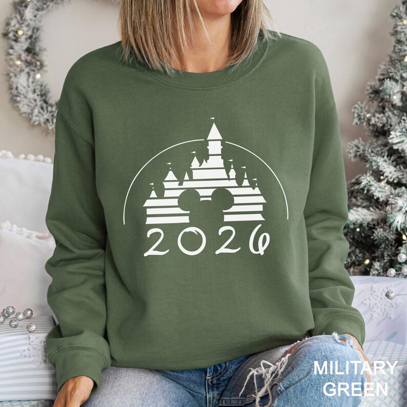 A woman wearing a green sweatshirt with a white graphic of a castle and the year "2026" printed on it.
