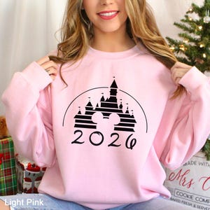A woman wearing a pink sweatshirt with a black graphic of a castle and the year "2026" printed on it.