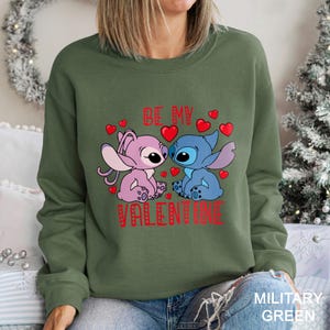 A woman wearing a green sweatshirt with a graphic design featuring two cartoon characters, one pink and one blue, along with the text "Be my Valentine".