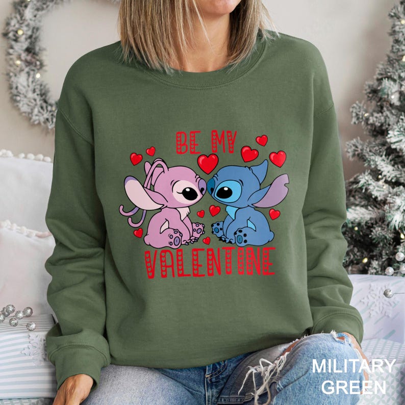 A woman wearing a green sweatshirt with a graphic design featuring two cartoon characters, one pink and one blue, along with the text "Be my Valentine".
