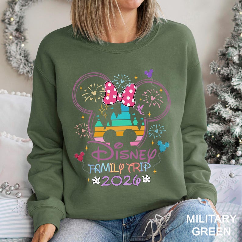 A woman wearing a green sweatshirt with a Disney-themed design, including a Mickey Mouse ear and fireworks, is sitting in front of a Christmas tree.