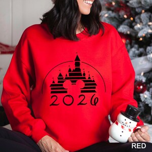 A woman wearing a red sweater with a black graphic of a castle and the year "2026" printed on it, holding a snowman toy.