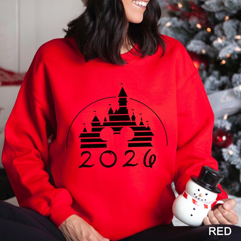 A woman wearing a red sweater with a black graphic of a castle and the year "2026" printed on it, holding a snowman toy.