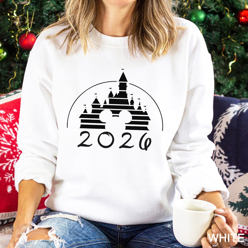 A woman wearing a white sweater with a black graphic of a castle and the year "2026" printed on it, sitting on a couch with a mug in her hand.