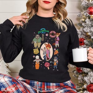 A woman wearing a black sweatshirt with a graphic of various cartoon characters from the animated film "Zooper 2" on it, holding a white mug.