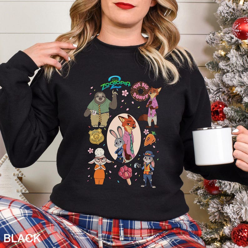 A woman wearing a black sweatshirt with a graphic of various cartoon characters from the animated film "Zooper 2" on it, holding a white mug.