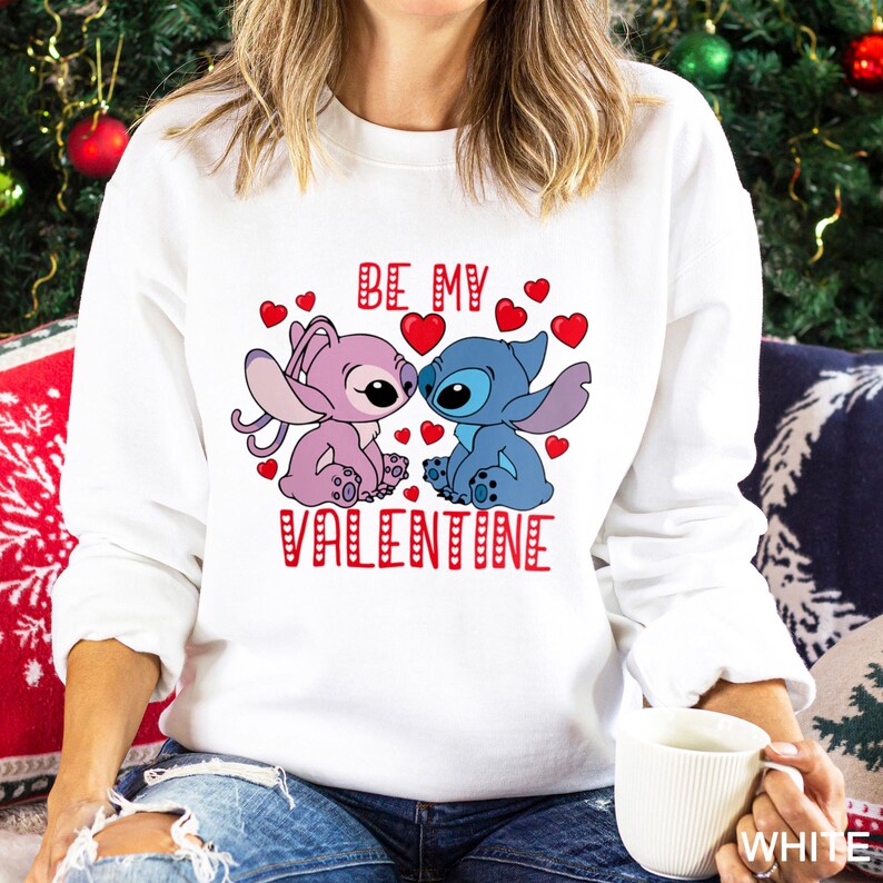A woman wearing a white sweatshirt with a graphic design featuring two cartoon characters, one pink and one blue, along with the text "Be my Valentine".