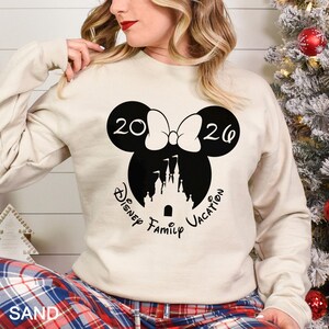 A woman wearing a white sweatshirt with a graphic design featuring a Minnie Mouse and the text "2020 Disney Family Vacation".