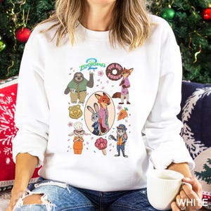 A woman wearing a white sweatshirt with a graphic of various cartoon characters from the movie "Zooper" printed on it. She is sitting on a couch and holding a white mug.