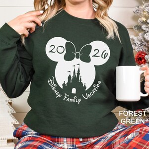 A woman wearing a green sweater with a white Mickey Mouse design and the text "Disney Family Vacation" printed on it, holding a white mug.