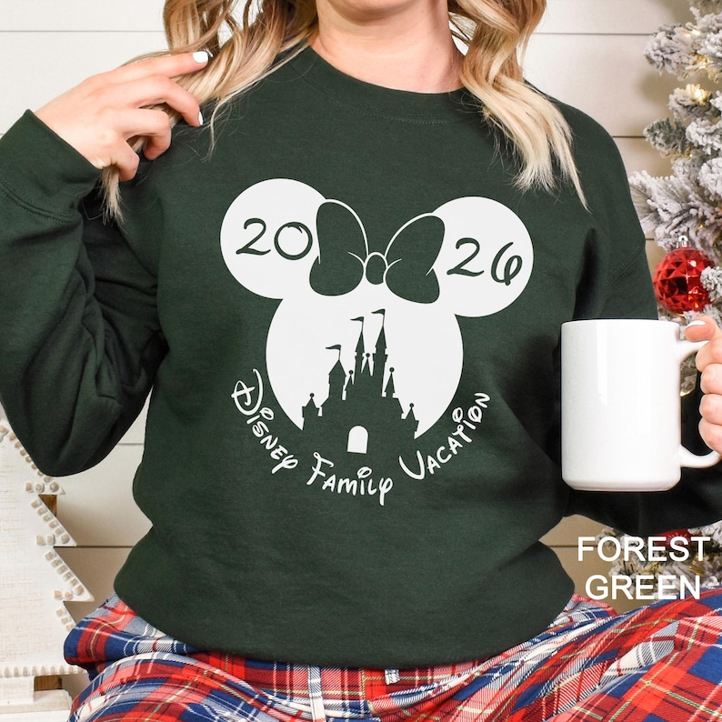 A woman wearing a green sweater with a white Mickey Mouse design and the text "Disney Family Vacation" printed on it, holding a white mug.