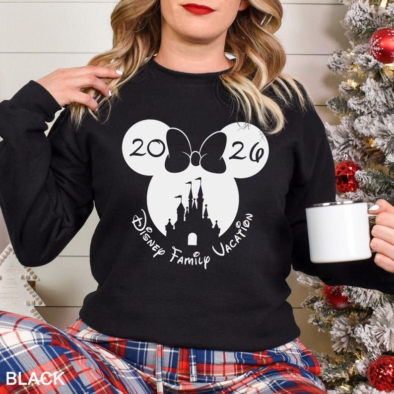 A woman wearing a black sweater with a white graphic design of a Mickey Mouse castle and the text "Disney Family Vacation" is holding a white mug.