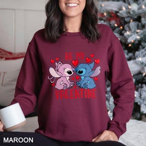 A woman wearing a maroon sweater with a graphic design featuring two cartoon characters, one pink and one blue, surrounded by hearts.