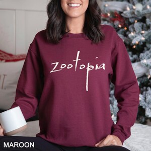 A woman wearing a maroon sweater with the word "Zootopia" printed on it, smiling and holding a mug.