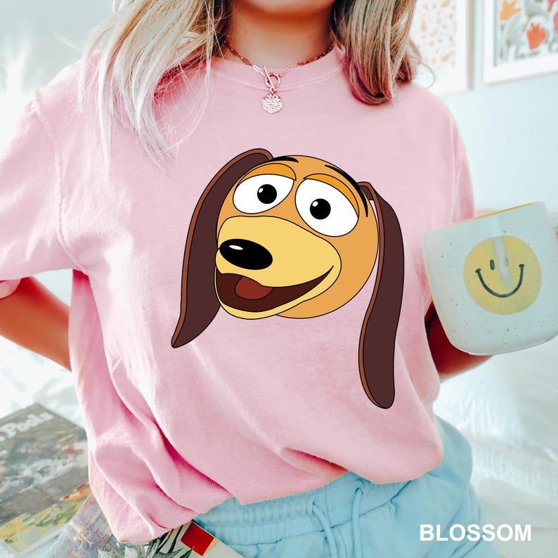 Puede incluir: Una camiseta rosa con una imagen de dibujos animados de un perro marr&oacute;n y amarillo con una gran sonrisa. El perro tiene una nariz marr&oacute;n y grandes ojos marrones.