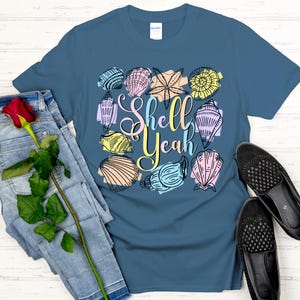 May include: A blue t-shirt with a colorful seashell design and the words "Shell Yeah". The design includes various seashell illustrations in pastel colors. A pair of black shoes and blue jeans are next to the shirt.