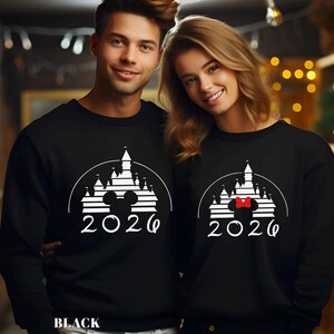 A man and a woman are standing next to each other, both wearing black sweatshirts with a white graphic design featuring a castle and the year "2020".