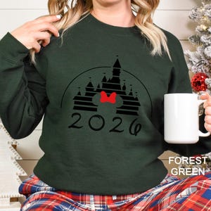 A woman wearing a green sweater with a graphic of a castle and the year "2026" printed on it, holding a white mug.