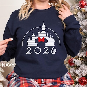 A woman wearing a navy blue sweatshirt with a white and red graphic of a Disney castle and the year "2026" printed on it.