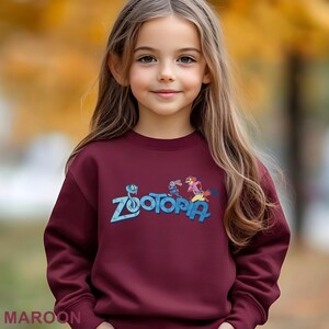 A young girl with long blonde hair wearing a maroon sweater with the word "Zootopia" printed on it.