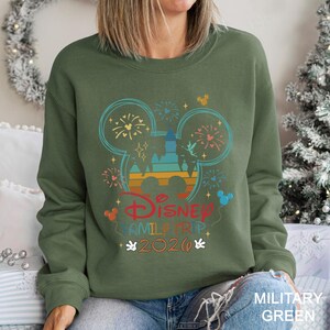A woman wearing a green sweatshirt with a Disney-themed graphic and text, sitting in front of a Christmas tree.