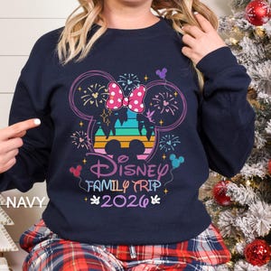 A woman wearing a navy blue sweatshirt with a Disney-themed design, including a Mickey Mouse ear and fireworks, is posing in front of a Christmas tree.