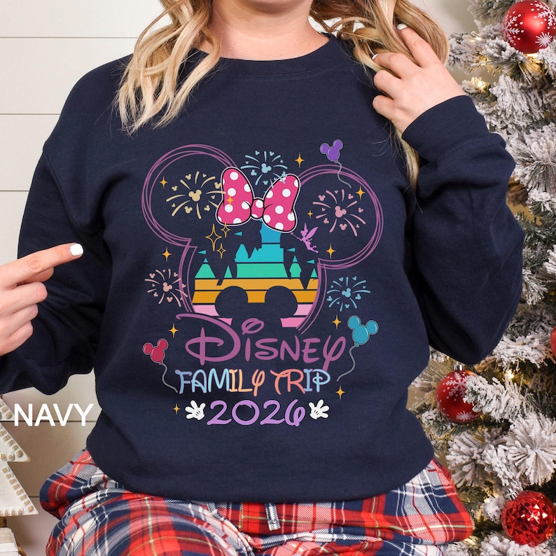 A woman wearing a navy blue sweatshirt with a Disney-themed design, including a Mickey Mouse ear and fireworks, is posing in front of a Christmas tree.