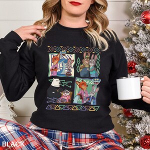 A woman wearing a black sweater with a colorful graphic of cartoon characters is holding a white mug.